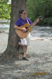 Me leaning against a tree with guitar in hand near a river.